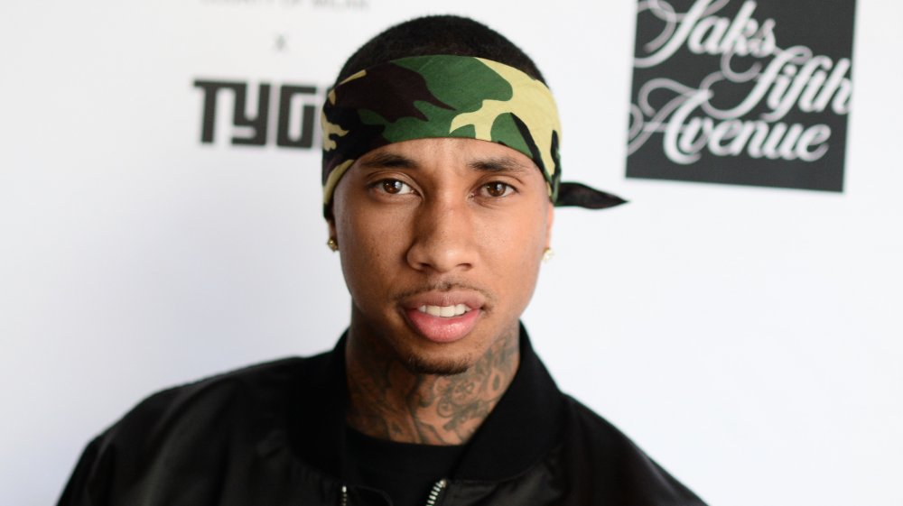 Tyga wearing a black bomber jacket with a camo bandanna, with an angry expression, looking straight at the camera