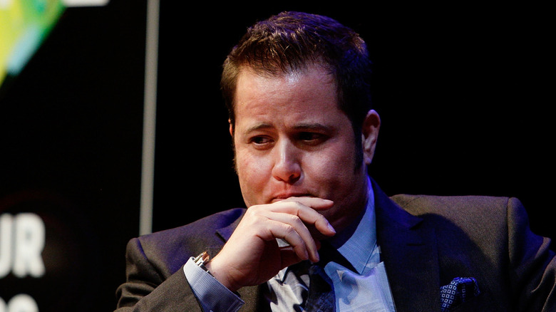 Chaz Bono wearing a suit and tie with his hand by his mouth during a speaking engagement