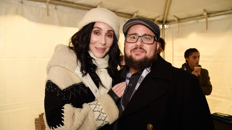 Cher wearing a tan beanie and tan and white scarf and winter jacket with Chaz Bono wearing a black pageboy cap and black winter jacket