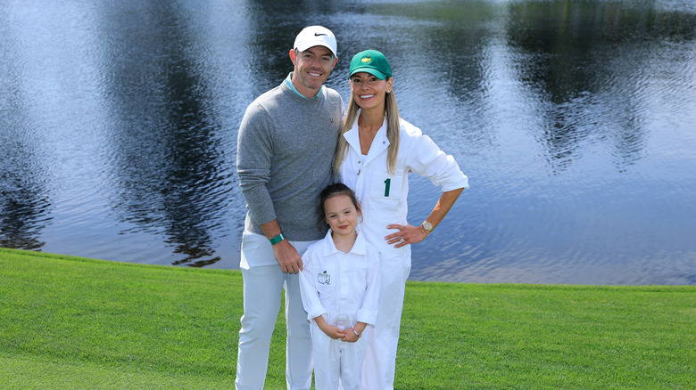 Rory McIlroy posing with his wife and daughter in front of a pond on a golf course.