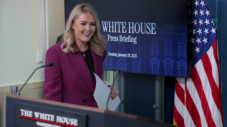 Karoline Leavitt smiling while approaching the White House press briefing room podium with papers in hand