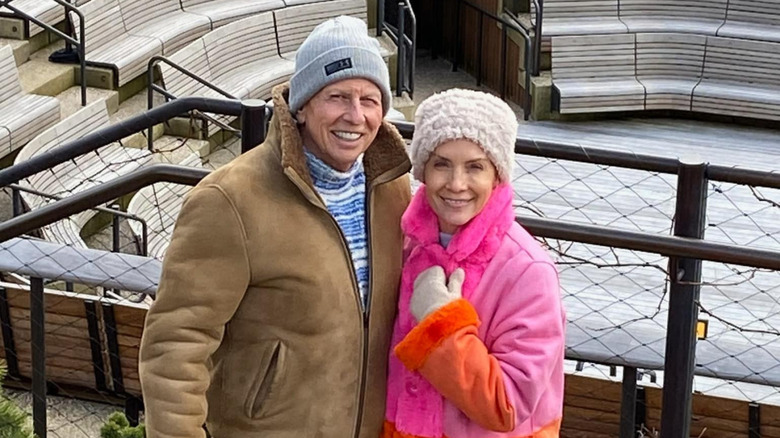 Dana Perino and Peter McMahon bundled up outdoors in front of a fence and seating.