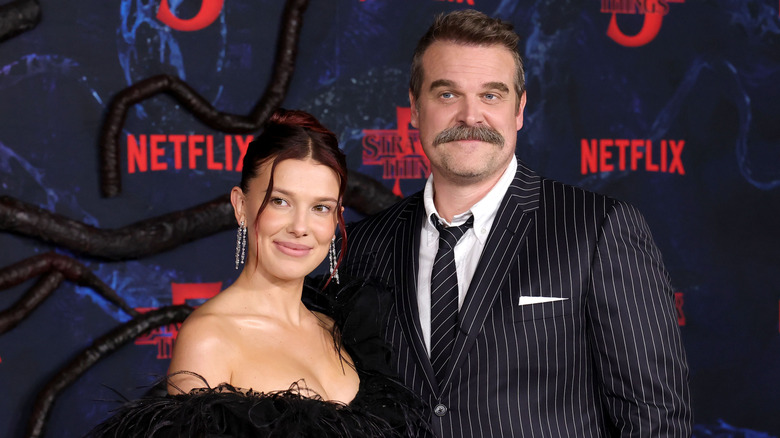 Millie Bobby Brown wearing a black lace and feather strapless gown beside David Harbour wearing a black and white pinstriped suit and tie