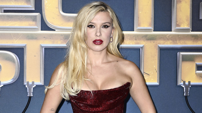 Millie Bobby Brown wearing a strapless, red gown with long, platinum blonde hair and red lipstick