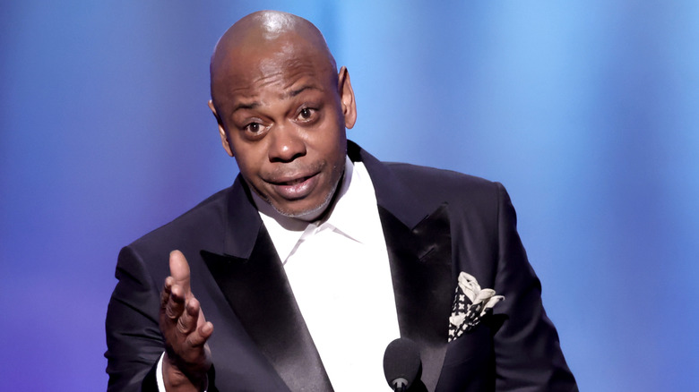 Dave Chappelle talking into a microphone with a hand up