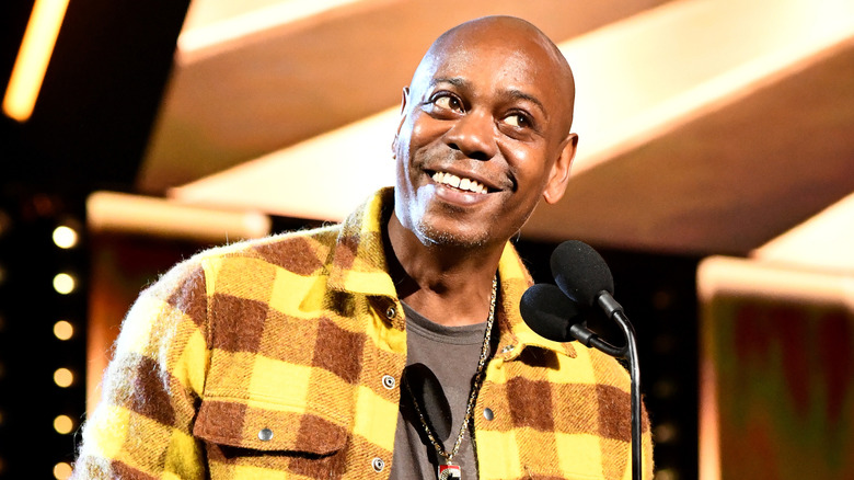 Dave Chappelle smiling and standing in front of a microphone.