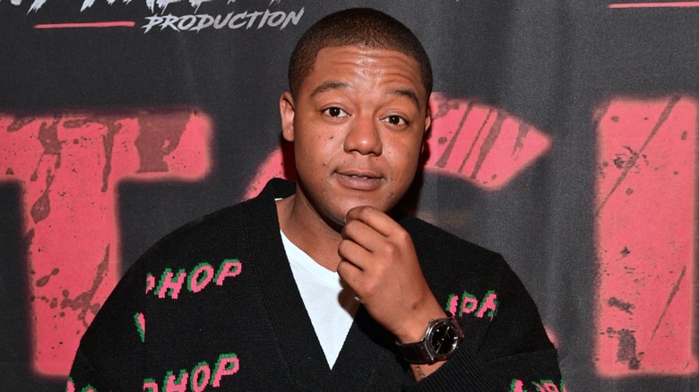 Sporting a black sweater with red detail, Kyle Massey raises his eyebrows while rubbing his chin.