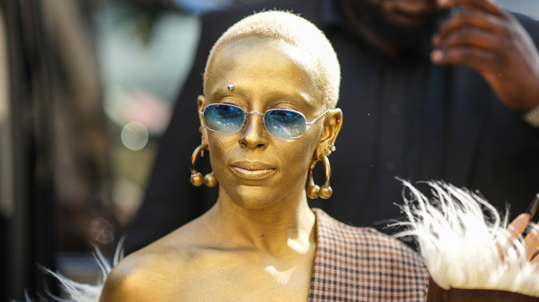 Doja Cat spray painted gold with gold dangling earrings, blue-tinted sunglasses, a tartan patterned shawl, and a shaved head
