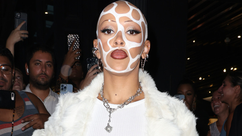 Doja Cat wearing a white fur coat, white ribbed top, long dangling silver necklace and earrings, and white netted face covering