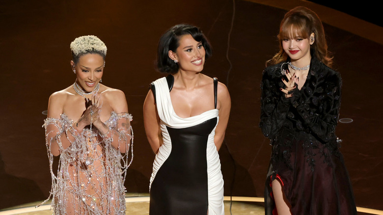 Doja Cat wearing a silver sheer draped gown with Raye wearing a black and white gown and Lisa wearing a black and maroon embellished long-sleeved gown