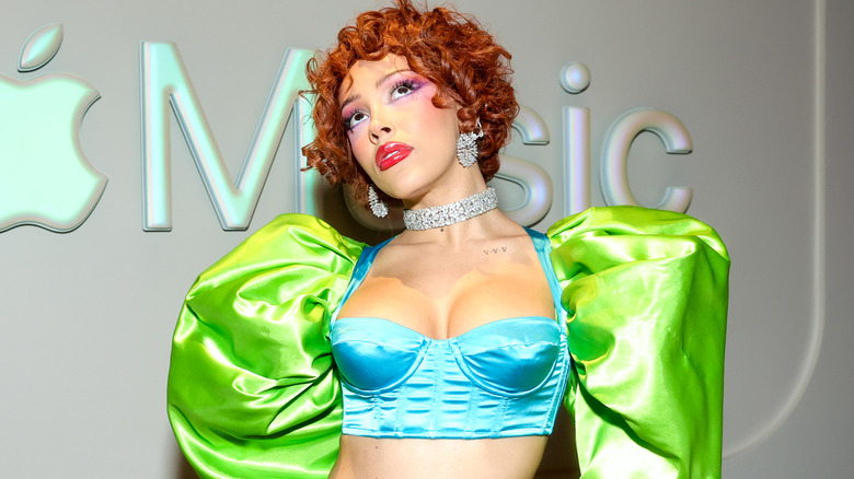 Doja Cat wearing a neon green and blue bustier top and puffy sleeves with red leggings, leopard print high waisted underwear, a sparkling choker necklace and earrings, and a red curly wig