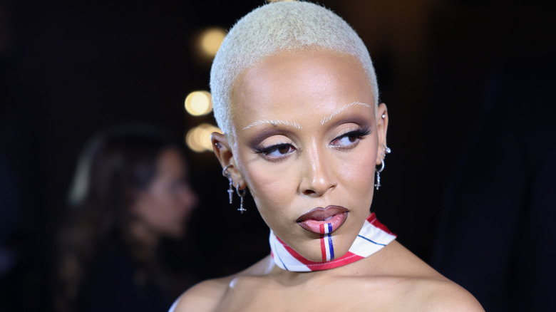 Doja Cat wearing red, blue, and white striped scarf, sparkling earrings and a bleached blonde shaved head and eyebrows