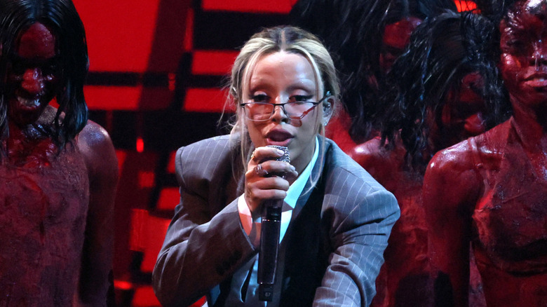 Doja Cat performing holding a microphone wearing a striped blazer, glasses, and collared shirt