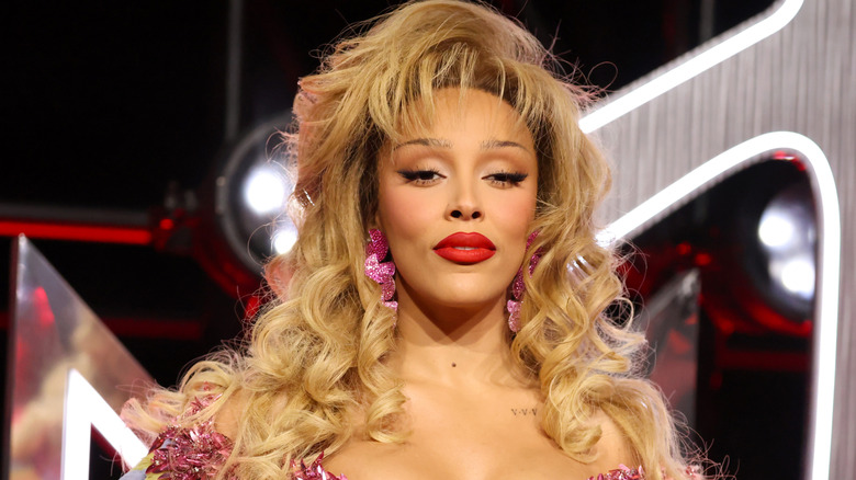 Doja Cat wearing an argyle patterned and feathered low cut dress with a lipstick shaped purse and feathered blonde wig