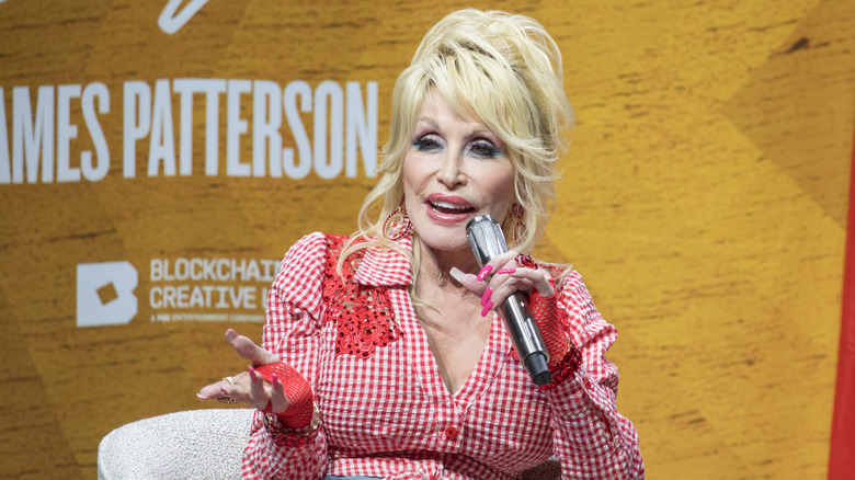 Dolly Parton speaking into a microphone in a red and white gingham dress onstage in 2022
