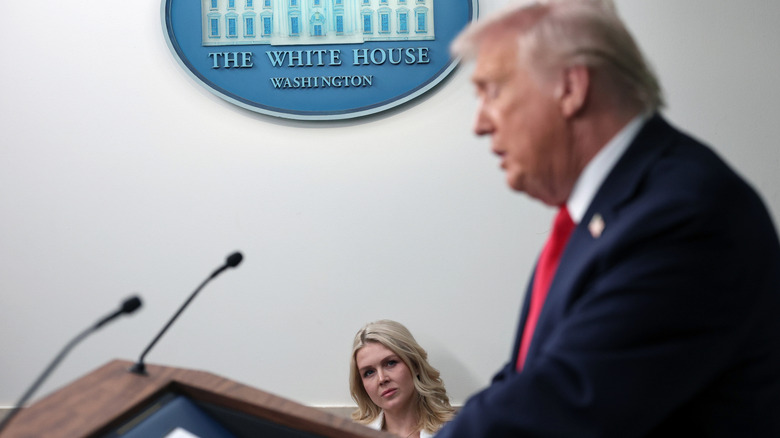 Karoline Leavitt watching Donald Trump speak at a lectern in the James S. Brady Press Briefing Room of the White House