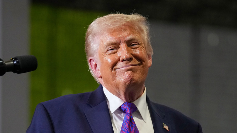 Donald Trump smiling in front of a microphone in a navy suit with a purple tie