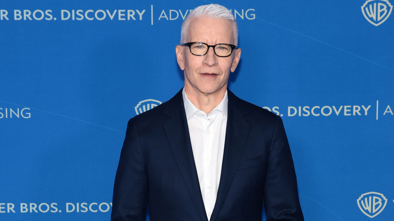 Anderson Cooper posing at an event in a black suit with an open white shirt and glasses