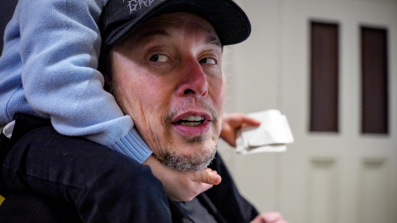 Elon Musk with a partially visible child on his shoulders, who is cupping the whiskers on Musk's chin