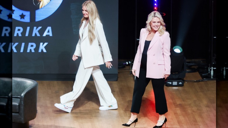 Erika Kirk wears a white suit and walks on stage beside Karoline Leavitt, who wears a pink blazer and black pants.