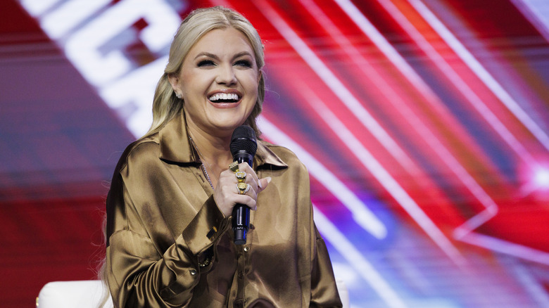 Erika Kirk wearing a gold outfit while smiling and holding a microphone to her mouth