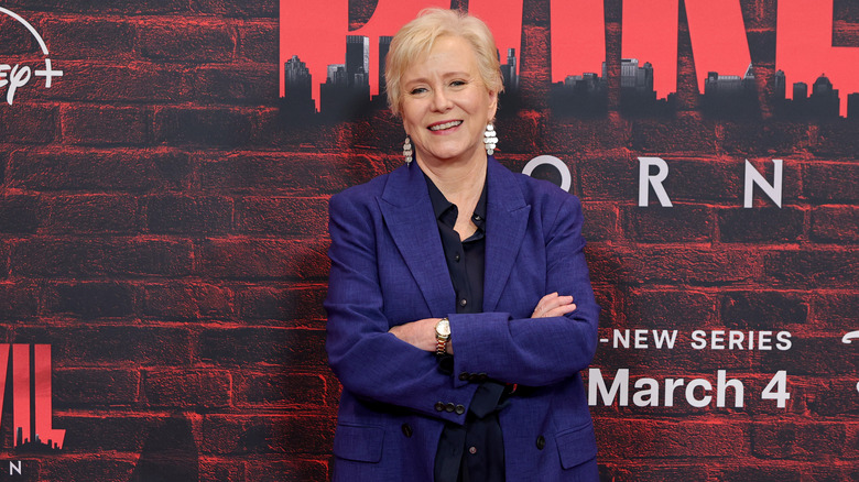 Eve Plumb smiling with her arms crossed and wearing a blue blazer at a red carpet event.