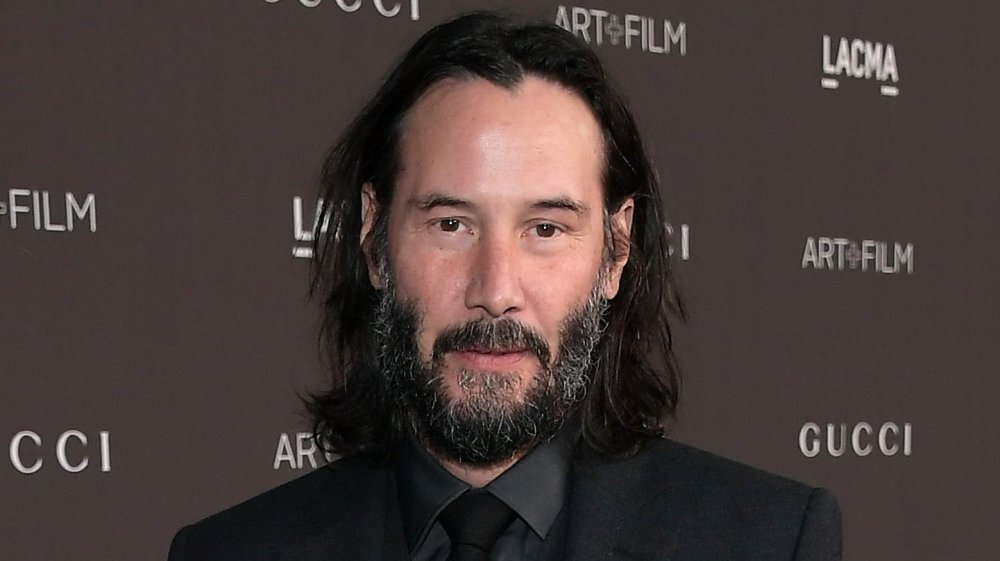 Keanu Reeves in a black three-piece suit, with longer hair tucked behind his ears, looking straight at the camera with a small smile