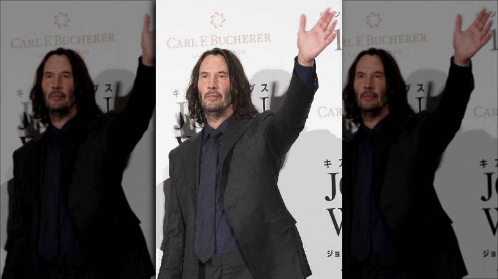 Keanu Reeves in a dark grey suit, with longer hair, waving at a red carpet event