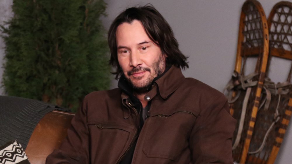 Keanu Reeves in a brown jacket, sitting and posing with a small smile at Sundance