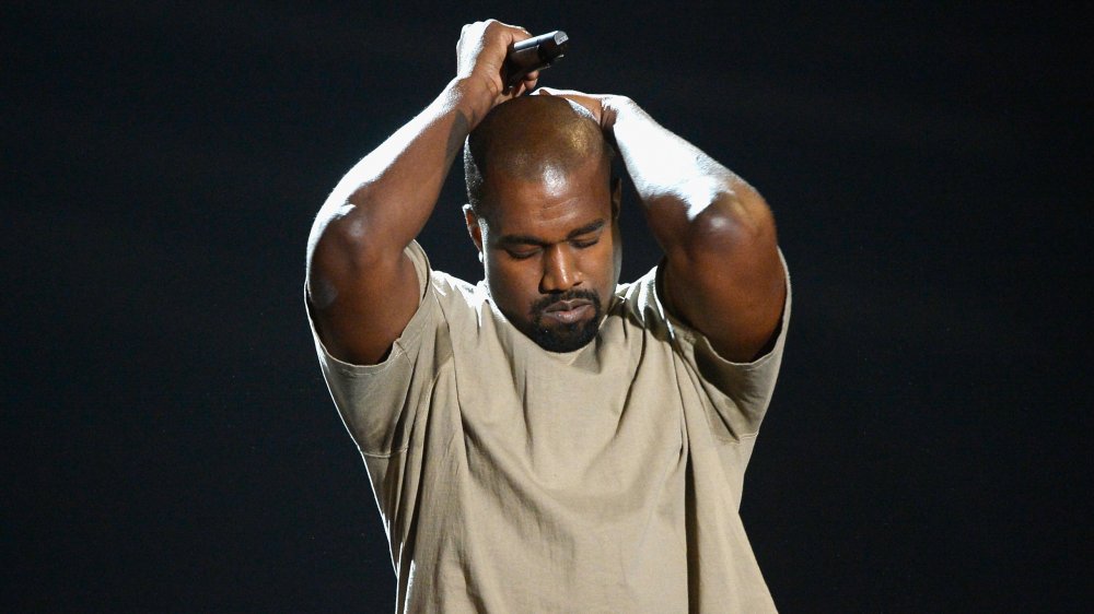 Kanye West in a beige t-shirt, holding his head with his eyes closed while on stage