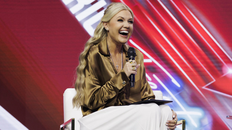Erika Kirk laughing on stage at America Fest 2025 in a brown silk shirt and white pants while holding a microphone