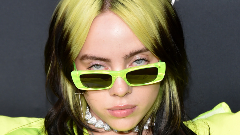Billie Eilish peering over a pair of green sunglasses with her hair dyed green and black