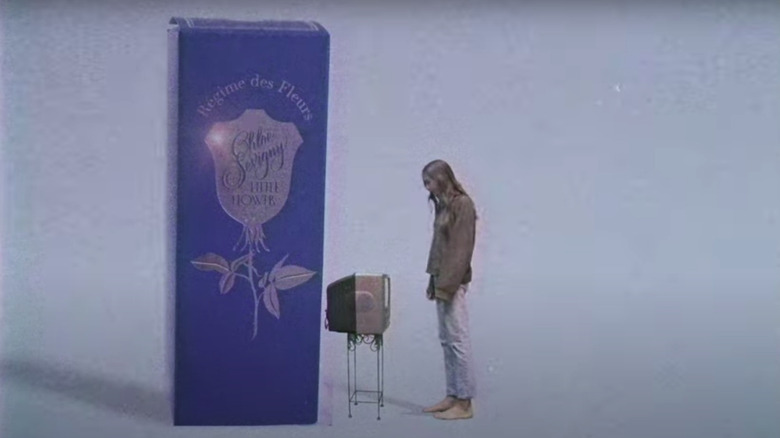 Chloe Sevigny staring at a TV in front of massive perfume bottle