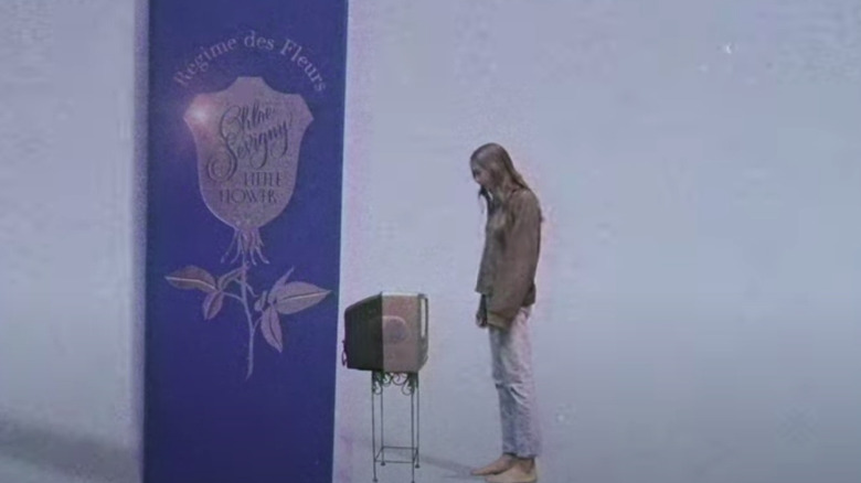 Chloe Sevigny staring at a TV in front of massive perfume bottle