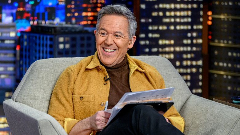 Greg Gutfeld smiling while sitting in chair holding papers