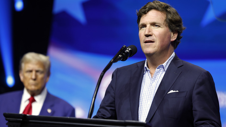 Tucker Carlson speaking on stage as Donald Trump looks on behind him.