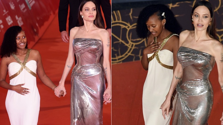 Zahara Jolie wears a white dress with gold straps crossing at the bust and Angelina Jolie wears a silver dress on the red carpet.