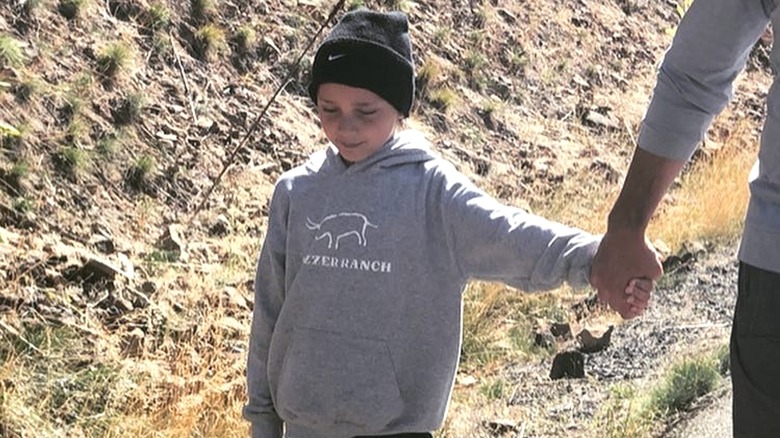 A young Duke Rancic wearing a gray sweatshirt looks downward while holding his father's hand