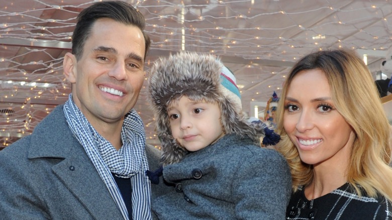 Young Duke Rancic, held between Bill and Guiliana Rancic gazes downward as his parents smile