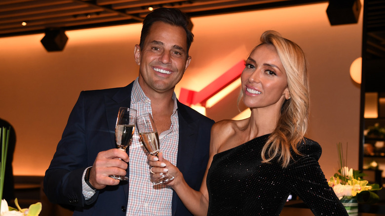 Bill and Giuliana Rancic holding champagne glasses wearing a black blazer and striped collared shirt and a black one-shouldered dress respectively