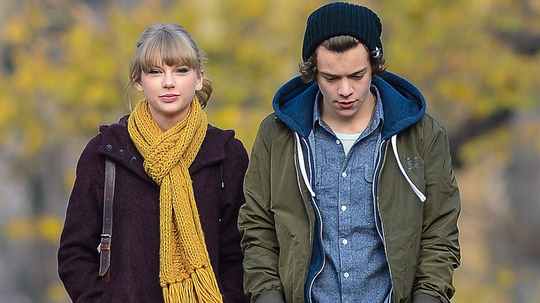 Taylor Swift wearing a dark jacket and yellow knit scarf with Harry Styles wearing a green hoodie, blue button-down shirt and black beanie
