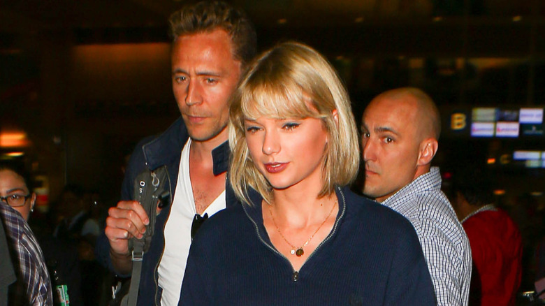Tom Hiddleston wearing a blue bomber jacket and white t-shirt with Taylor Swift wearing a blue blouse and gold necklace