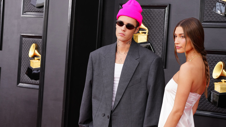 Justin Bieber wearing a pink beanie and charcoal gray suit jacket next to Hailey Bieber, who wears a white strapless dress.