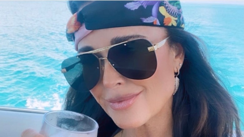 Kyle Richards, wearing sunglasses, smiling no teeth, wearing bandana, drink in her, ocean behind her