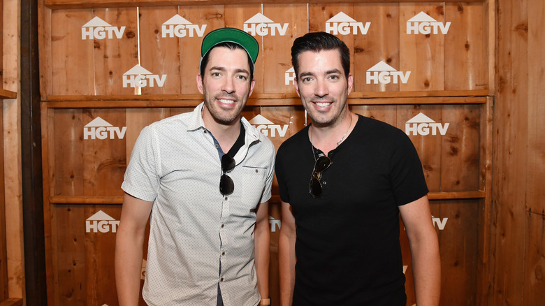 Drew and Jonathan Scott posing together in front of the HGTV logo.