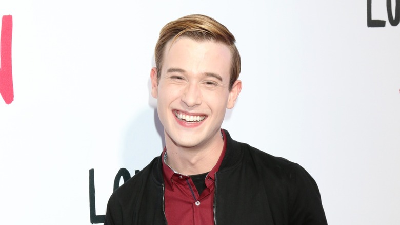 Dressed in a burgundy shirt and a black jacket Tyler Henry tilts his head and gives a toothy smile