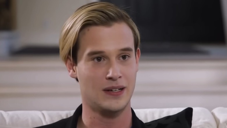 Sitting on a couch, Tyler Henry slightly parts his lips while looking away from the camera