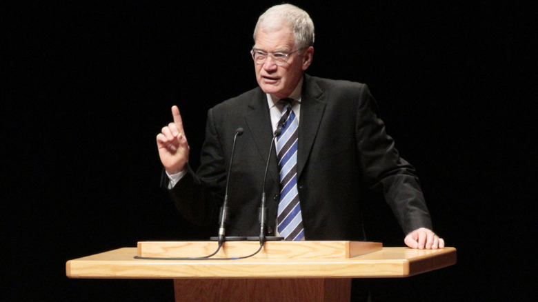 David Letterman holding up a finger while speaking on stage in a black suit