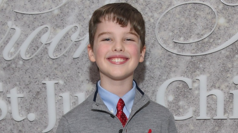 Dressed in a gray sweater, a blue shirt, and a maroon tie, Iain Armitage sports a wide smile.