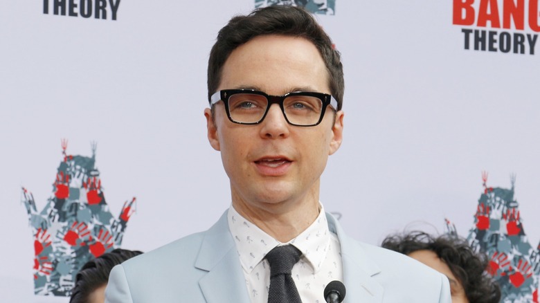 Wearing a set of glasses and a suit-tie combo, Jim Parsons speaks into a microphone.